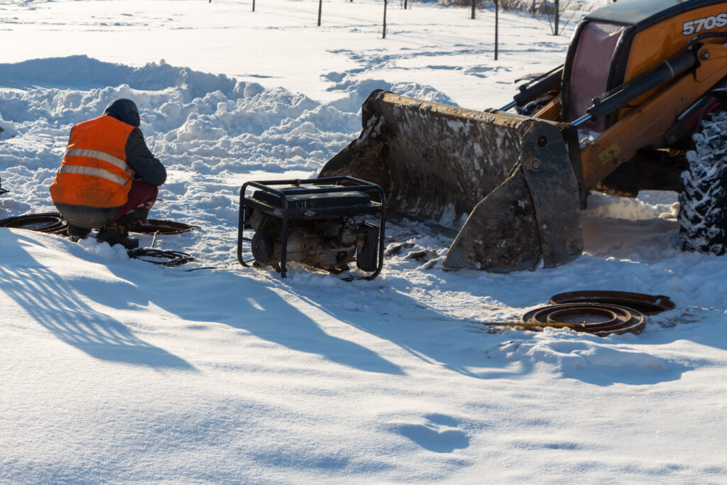 Electrician crouching in snow next to a generator and construction vehicle.