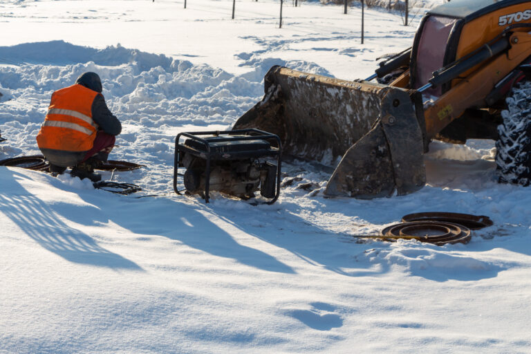 Electrician crouching in snow next to a generator and construction vehicle.